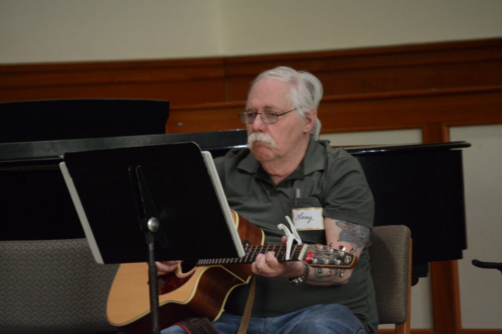 A white man with gray hair and a mustache plays an acoustic guitar while looking at a black music stand