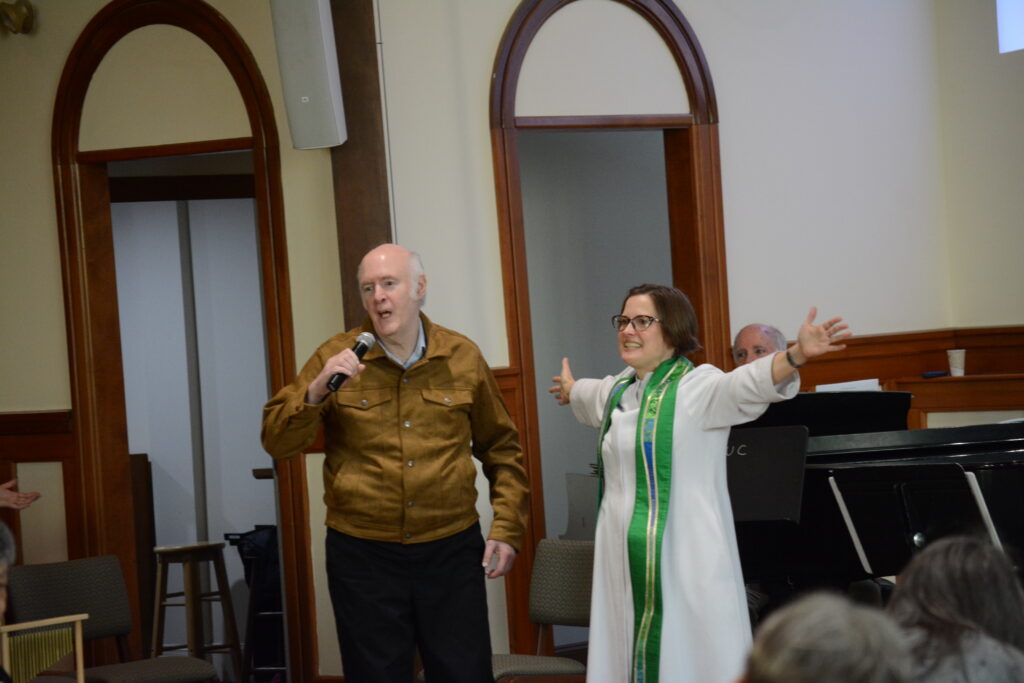 Pastor Hannah is wearing a white robe and Bruce is wearing a leather jacket and holding a microphone