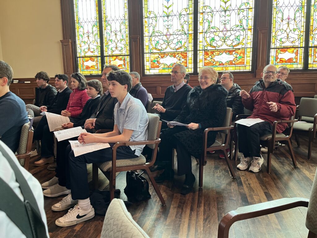 A group of people are seated in chairs in front of stained glass windows.