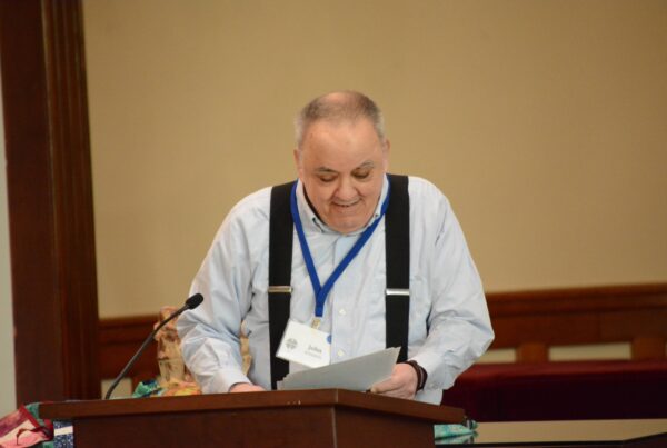 John Kilmartin speaks from a wooden podium. John is wearing a blue button up shirt and black suspenders.